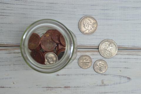 A photograph of a jar on a wooden table with coins in it and four coins scattered next to it.