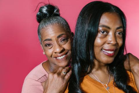 Two female presenting Black seniors smiling, behind them is a pink backdrop. 